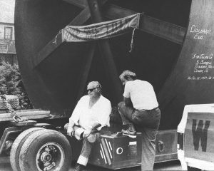 A old black and white photo featuring two men sitting in front of a oversize load.