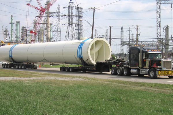A massive superload moves down the road in front of a series of electrical towers and powerlines.