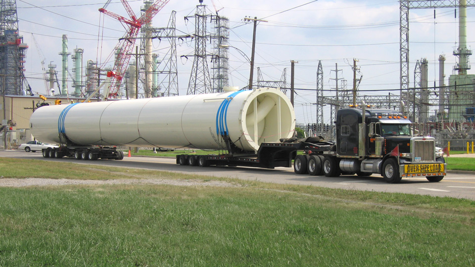 A massive superload moves down the road in front of a series of electrical towers and powerlines.