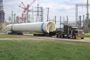 A massive superload moves down the road in front of a series of electrical towers and powerlines.