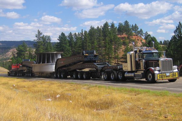 A large overside load moves uphill with blue cloudy skys, hills, and trees in the background.