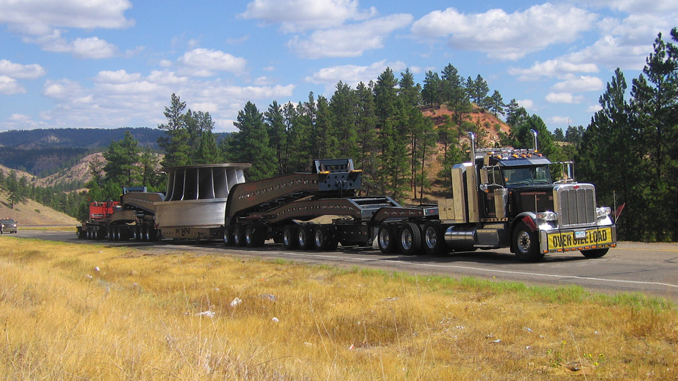 A large overside load moves uphill with blue cloudy skys, hills, and trees in the background.