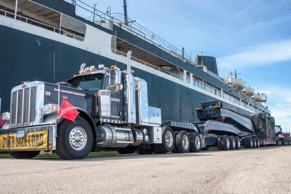 A 20 axle rig parked alongside a large ship.