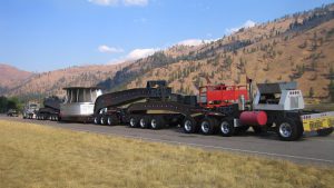 A massive superload moves down the road with hills and pine trees in the background.