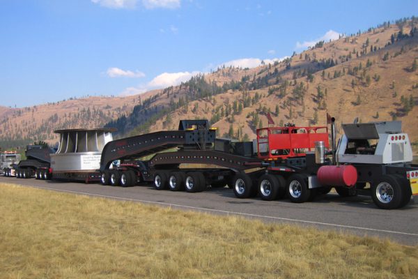 A massive superload moves down the road with hills and pine trees in the background.