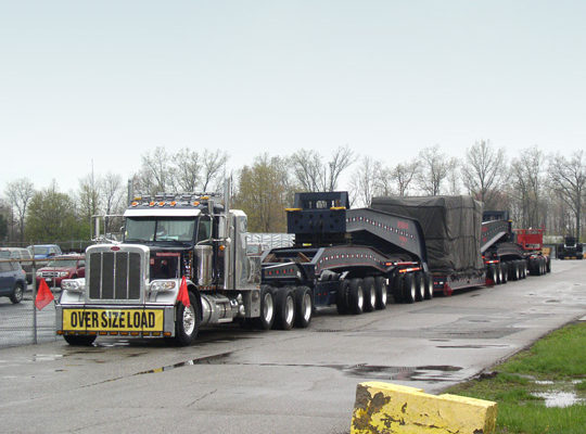 A 20 axle trailer, loaded with and injection mold machine clamp and sitting in a parking lot on an overcast day.