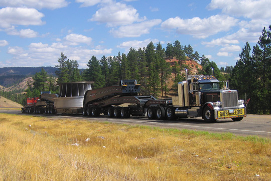 A large overside load moves uphill with blue cloudy skys, hills, and trees in the background.